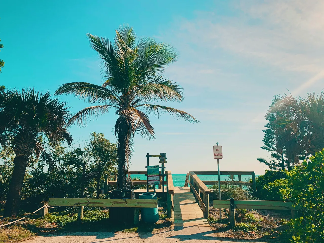 Palm-lined wooden beach path leading to turquoise ocean under a bright Florida sky.